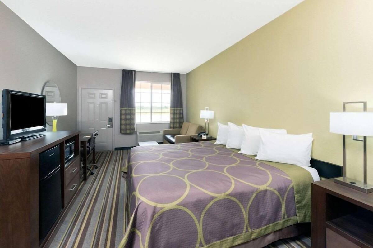 A hotel room with a bed, a television, a desk with a lamp, a chair, and a window with curtains.