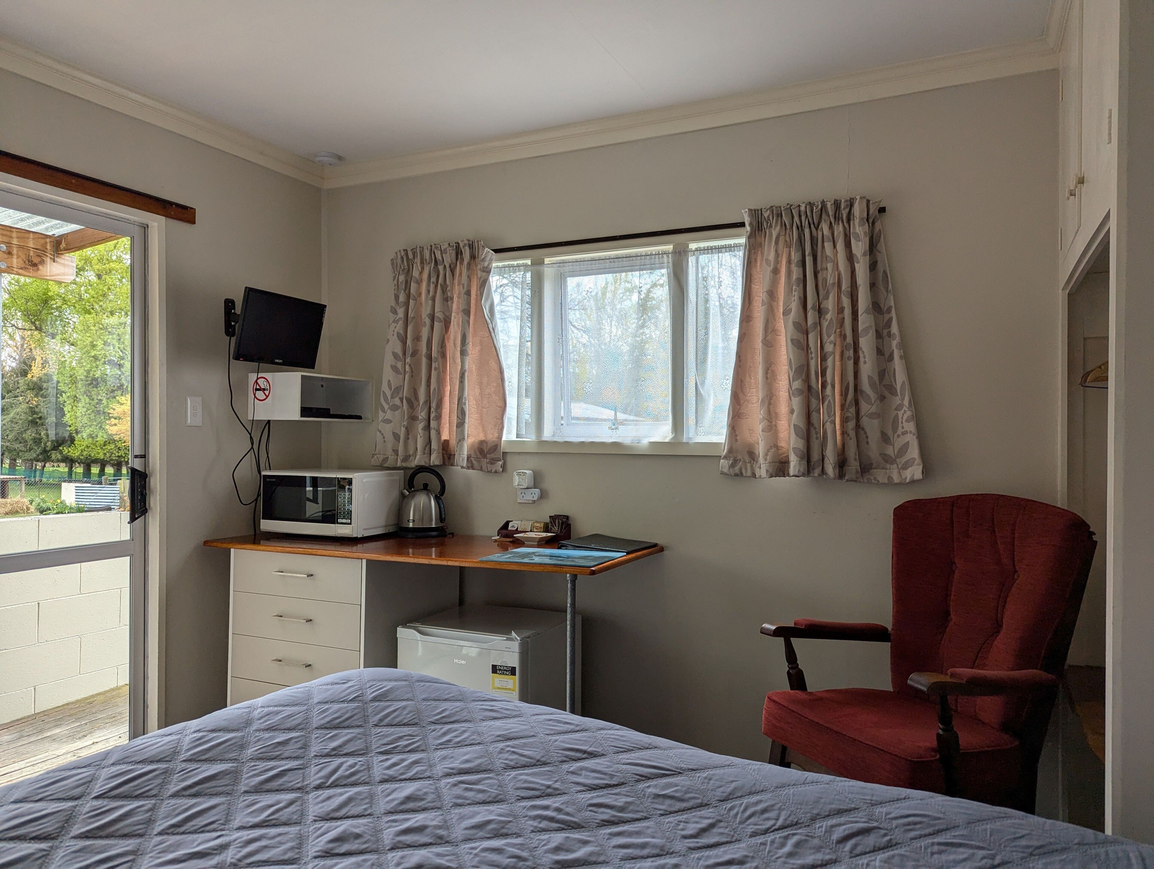 Een slaapkamer met een bed, een bureau met een computer, een stoel, een raam met gordijnen en uitzicht op groen buiten.