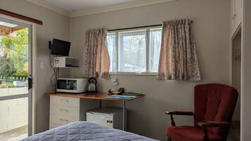 Een slaapkamer met een bed, een bureau met een computer, een stoel, een raam met gordijnen en uitzicht op groen buiten.