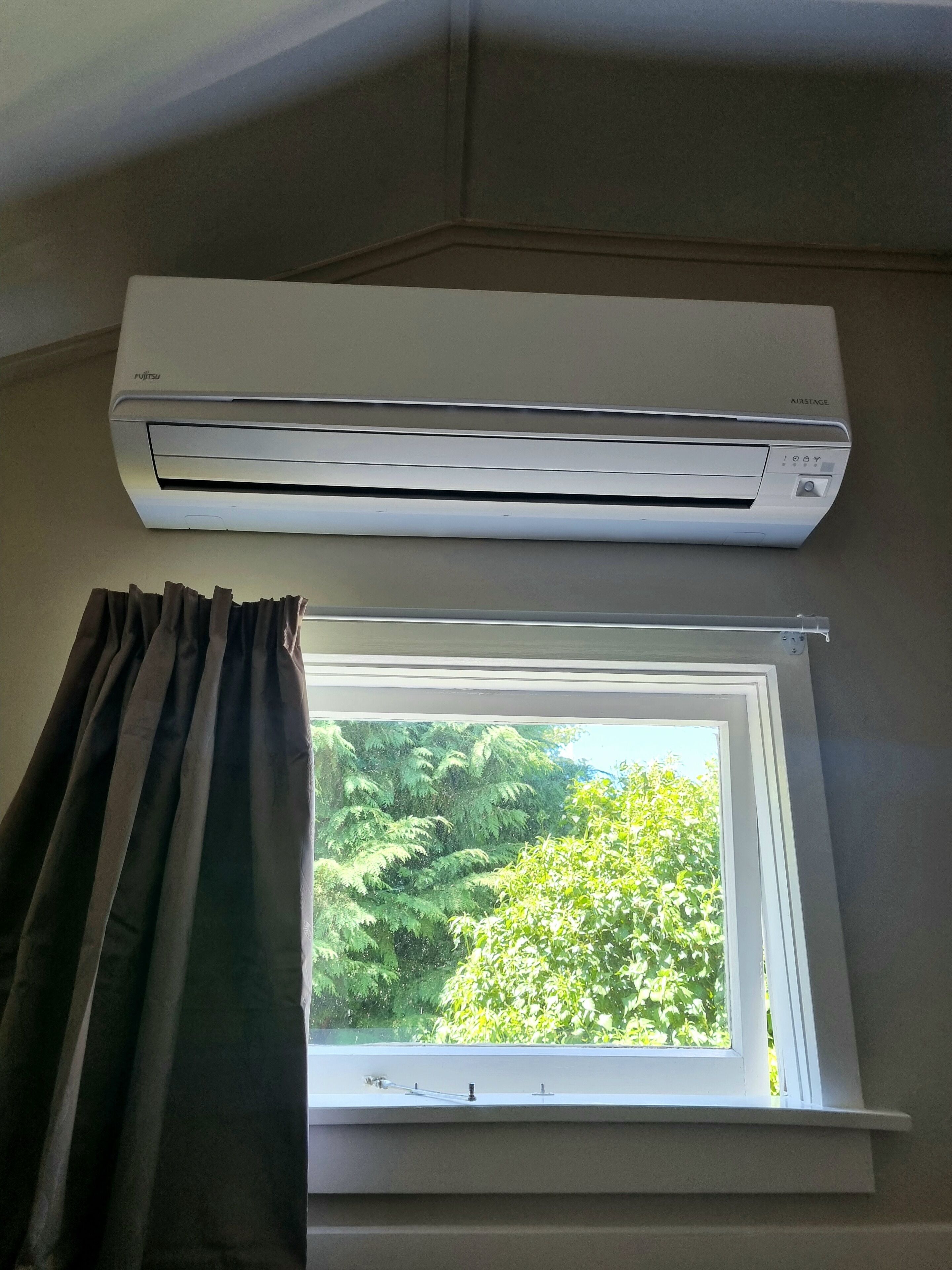 Une pièce dotée d’un climatiseur fixé au mur, au-dessus d’une fenêtre donnant sur un paysage verdoyant.