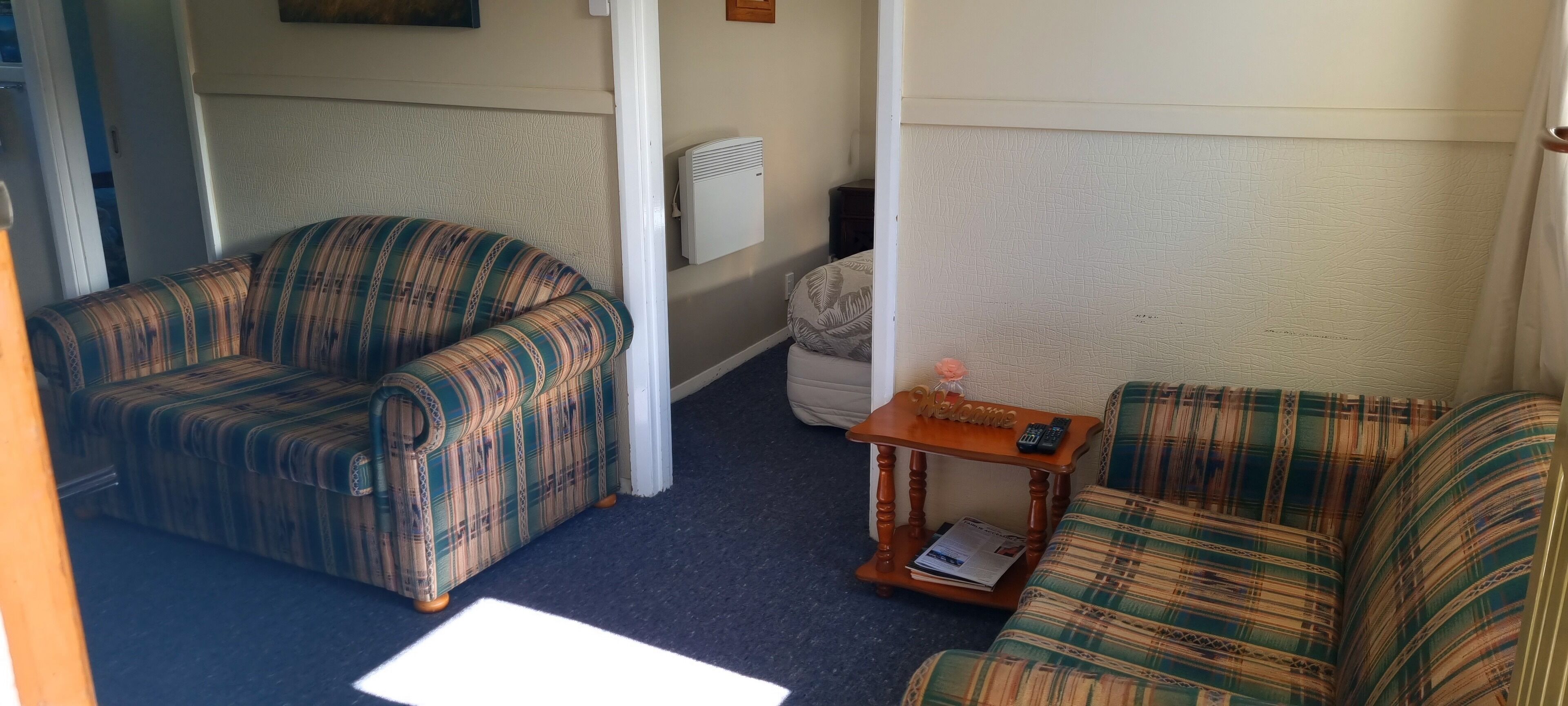 A living room with two patterned sofas, a small wooden side table with a remote control and a magazine, and a white radiator on the wall.