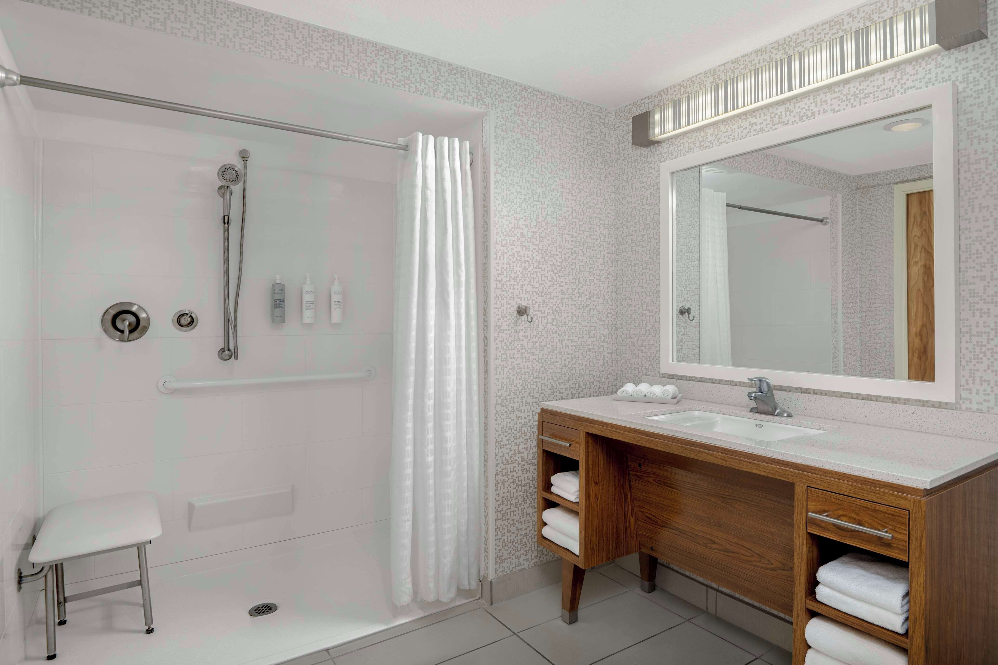 A modern bathroom with a glass-enclosed shower, a wooden vanity with a large mirror, and a small stool.