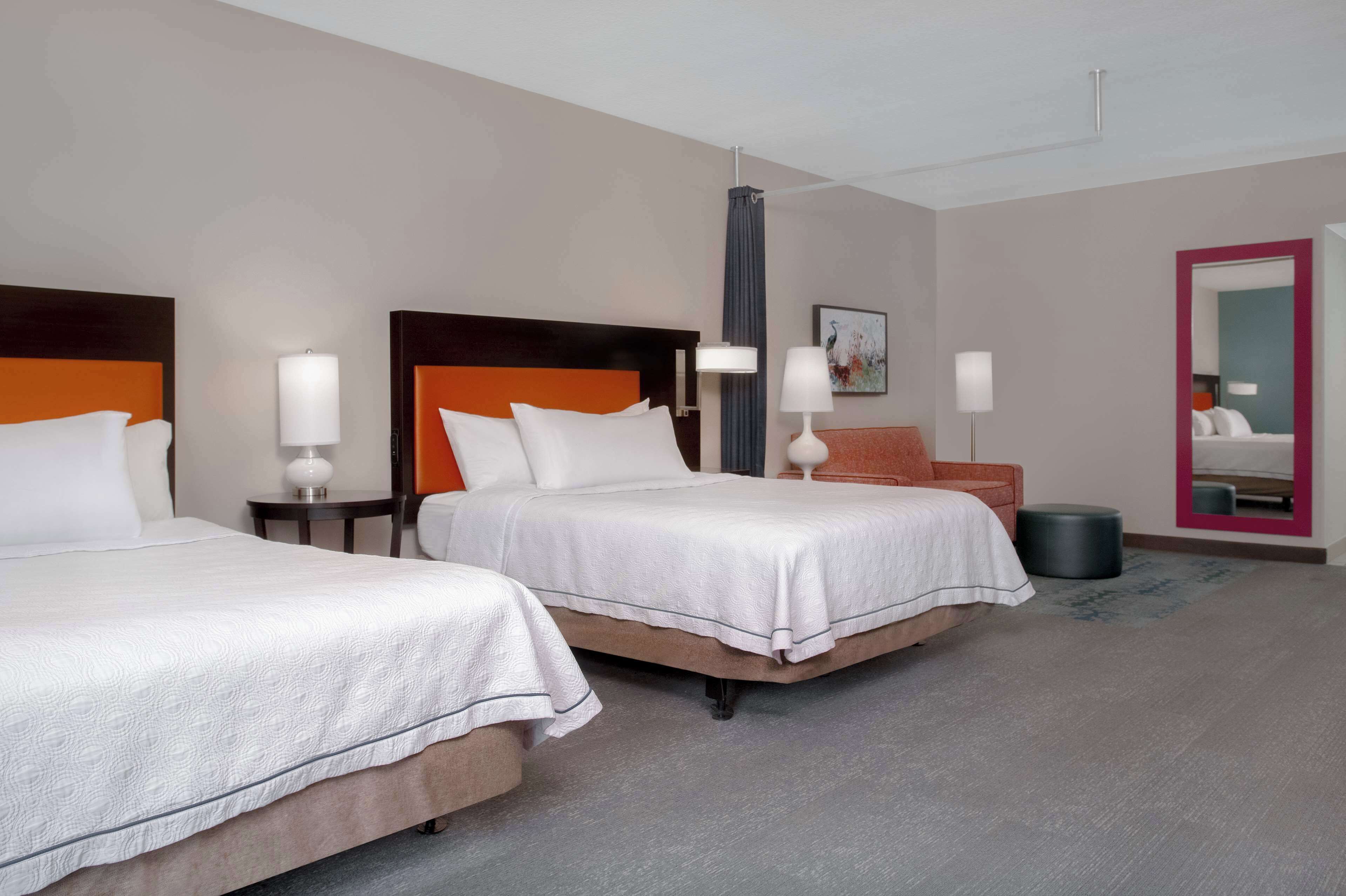 A hotel room with two beds, a nightstand, a lamp, a mirror, and a painting on the wall.