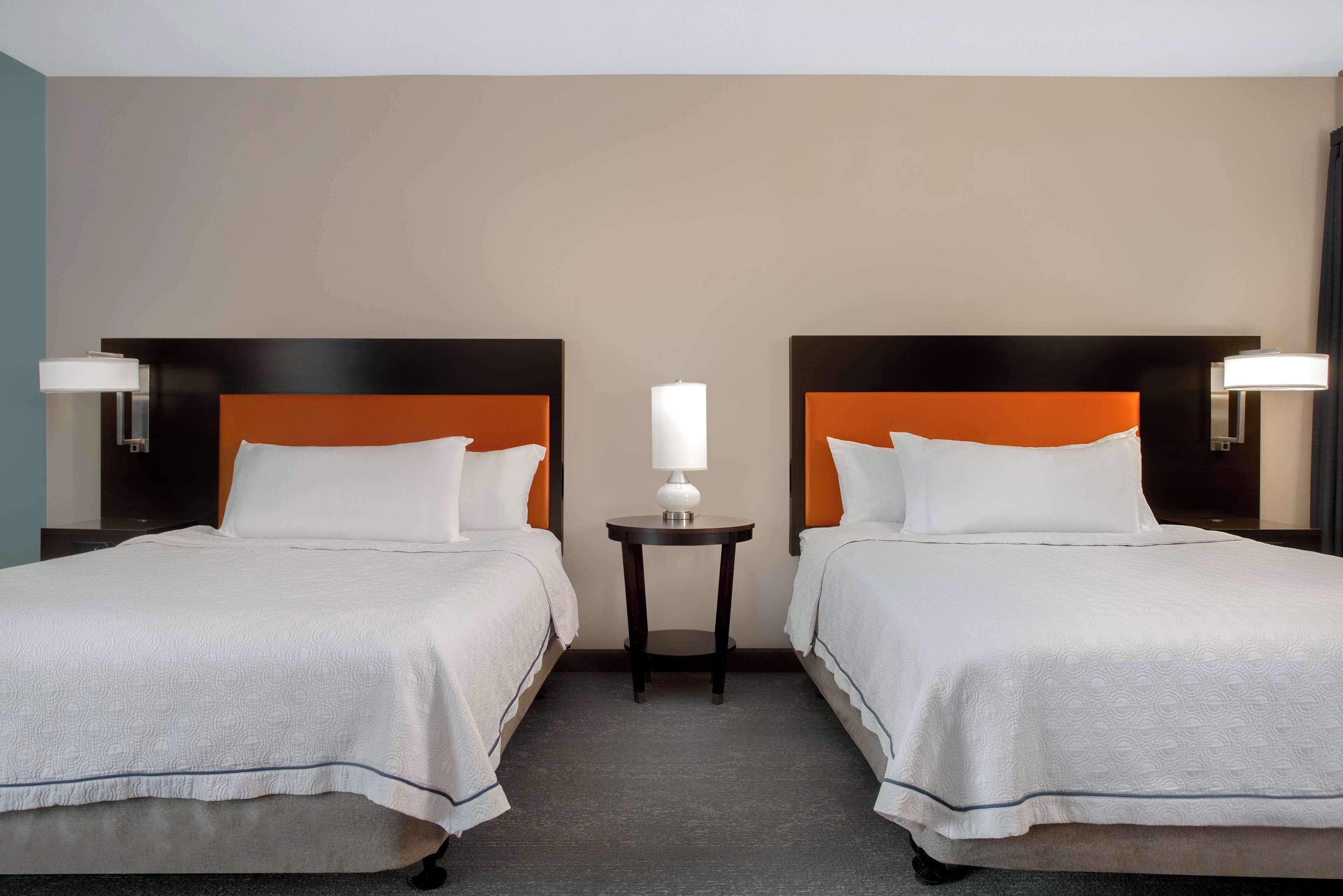 Une chambre d’hôtel avec deux lits, chacun doté d’une tête de lit orange, une petite table ronde avec une lampe et une moquette de couleur neutre.