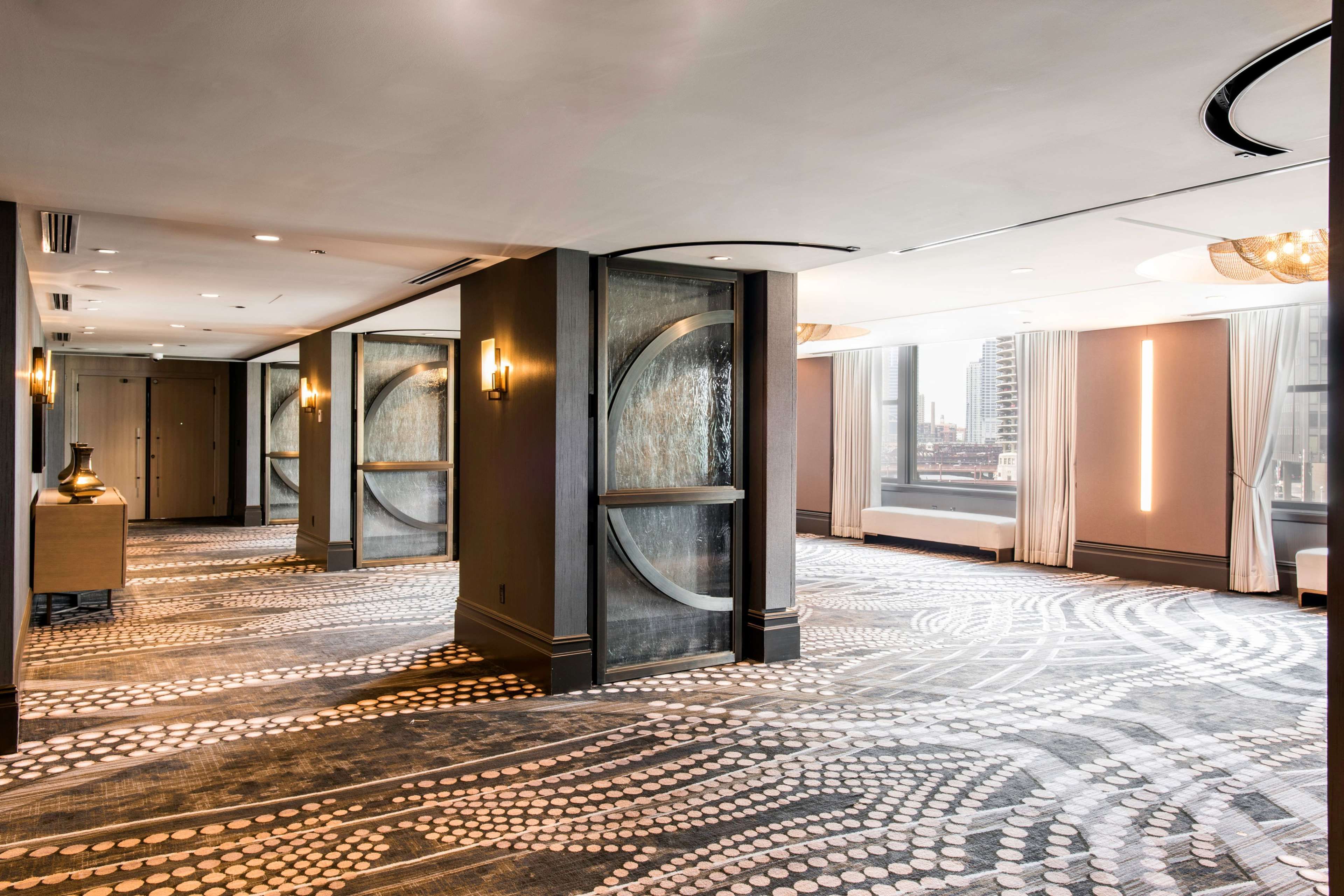 A hotel corridor with a patterned carpet, wall sconces, and a view of the city through large windows.
