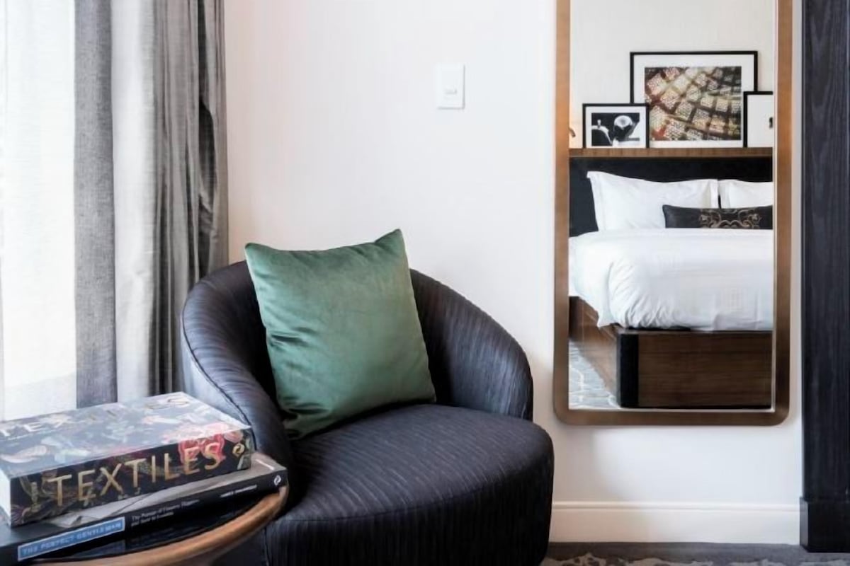 Camera d'albergo con una poltrona in pelle scura, un comodino con un libro intitolato “TEXTILES”, uno specchio grande che riflette un letto con biancheria bianca e quadri appesi al muro.