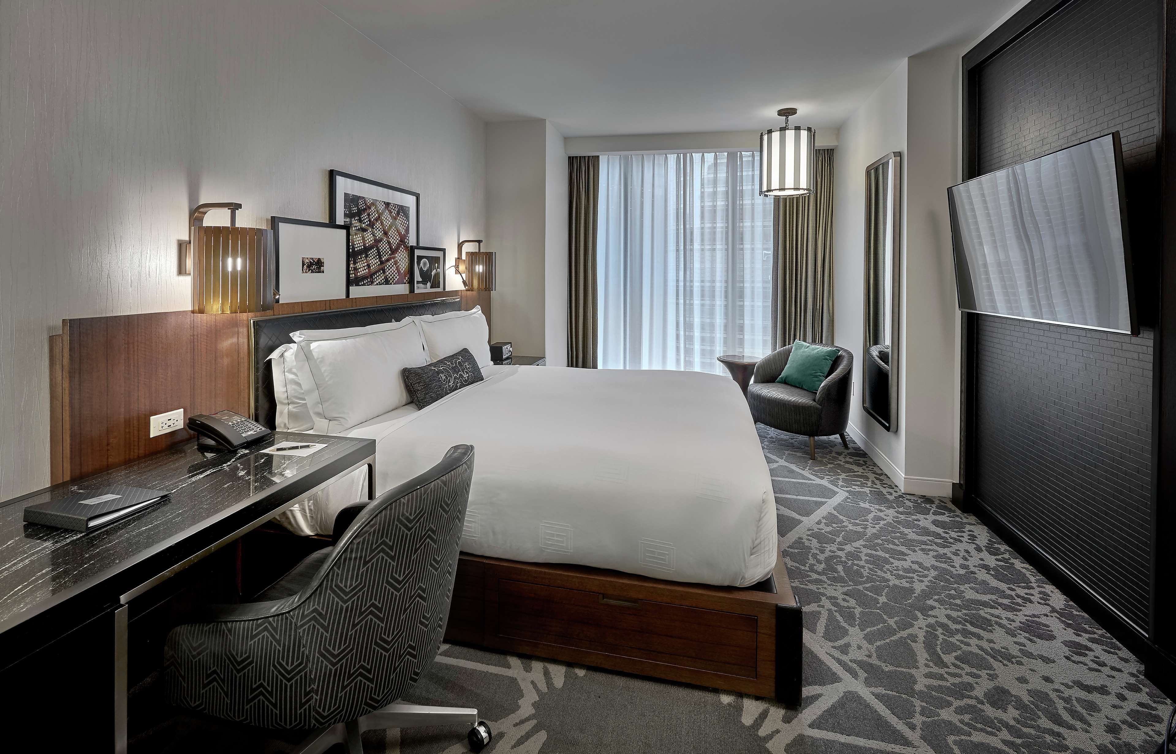 Quarto de hotel com cama, escrivaninha, cadeira e uma janela grande com cortinas.