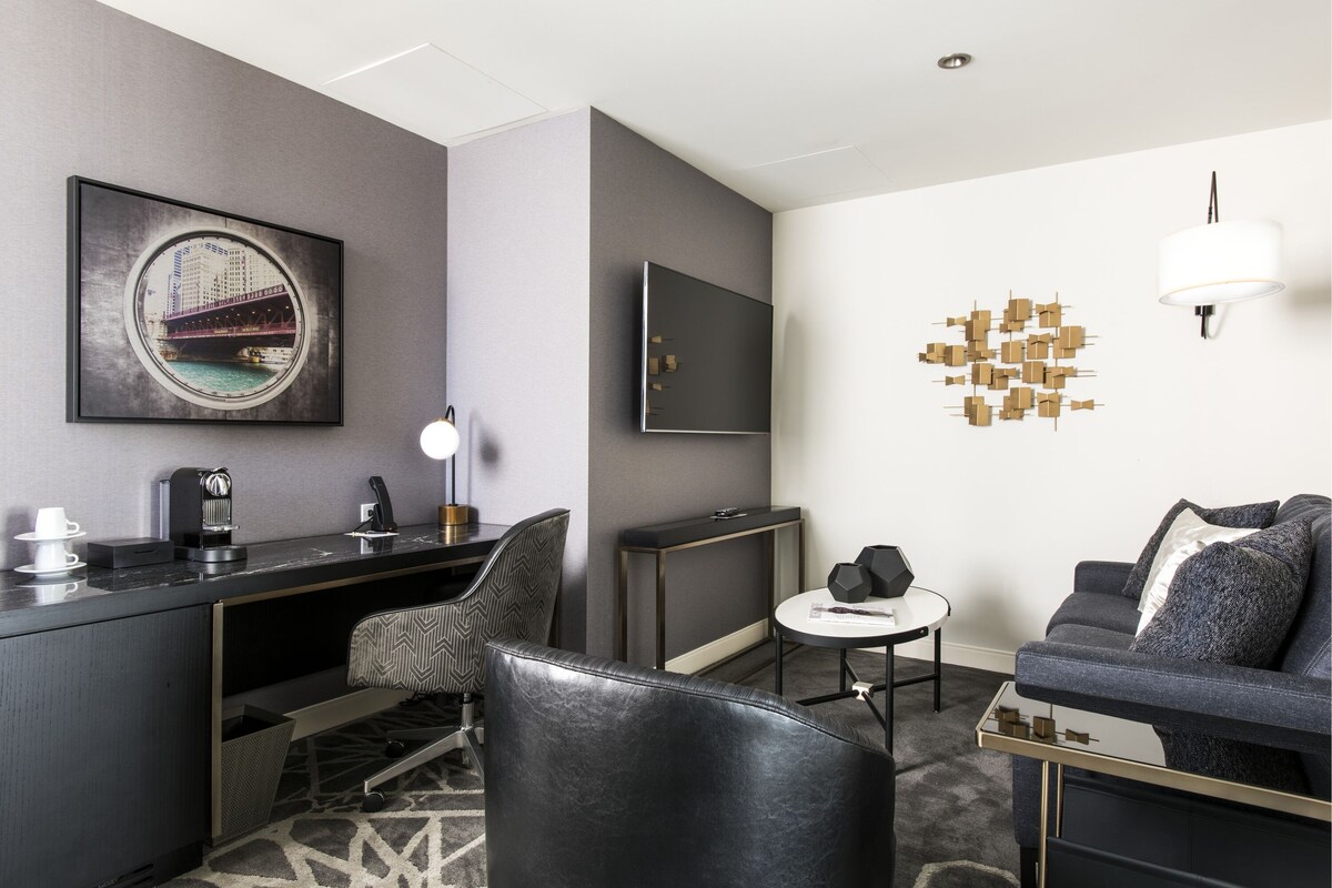A modern hotel room with a desk, a sofa, a coffee machine, a TV, and a unique wall art piece.