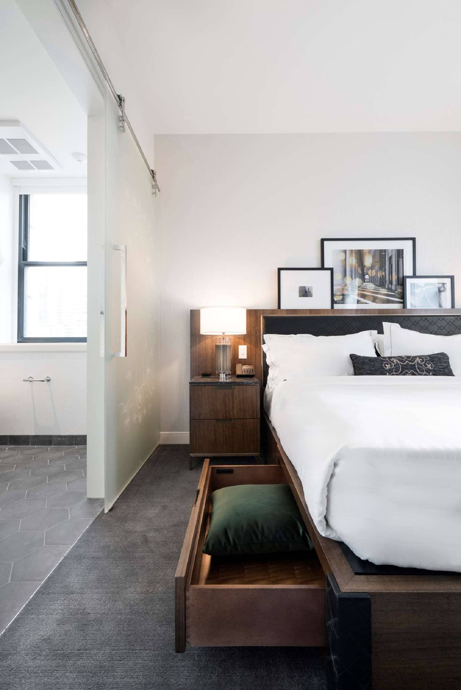 A hotel room with a bed, a nightstand with a lamp, a wooden drawer, and framed pictures on the wall.