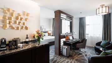 A hotel room with a marble countertop, a bed, a desk with a laptop, a chair, a sofa, a coffee machine, a wine bottle, and a decorative wall piece.