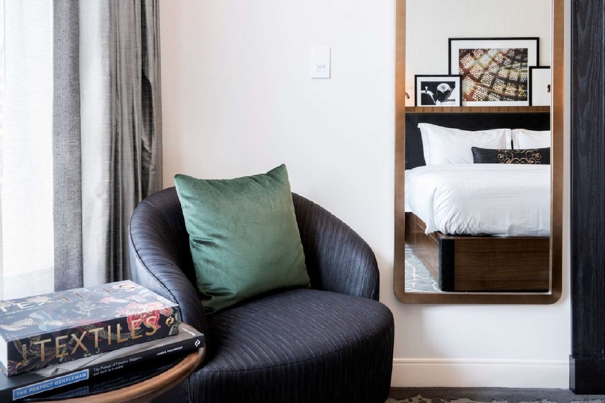 A hotel room with a black leather armchair, a small side table with books, a large mirror reflecting a neatly made bed, and a carpeted floor.