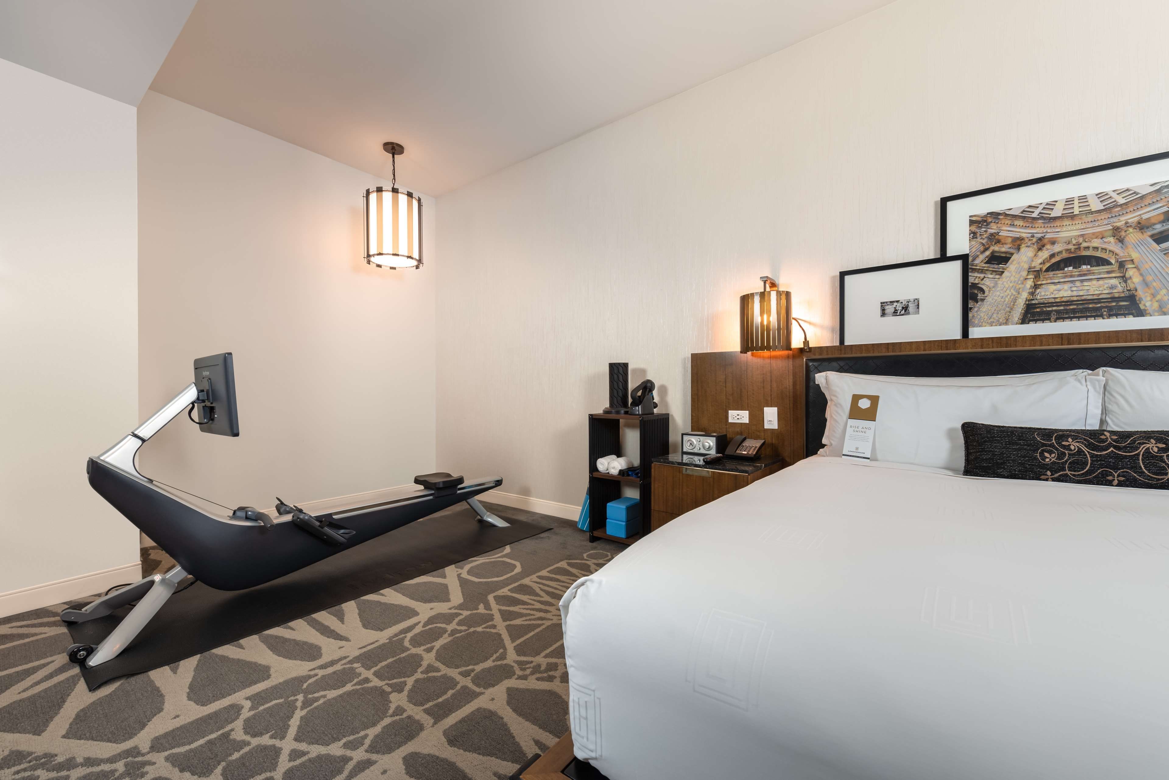 Habitación de hotel con cama, escritorio, silla, cinta de correr, televisor de pared y un cuadro con la imagen de un edificio.