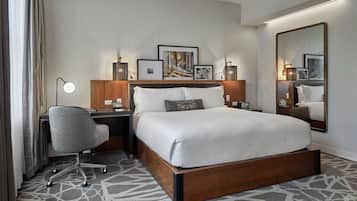A hotel room with a large bed, a desk with a chair, a desk lamp, framed pictures on the wall, and a large mirror.