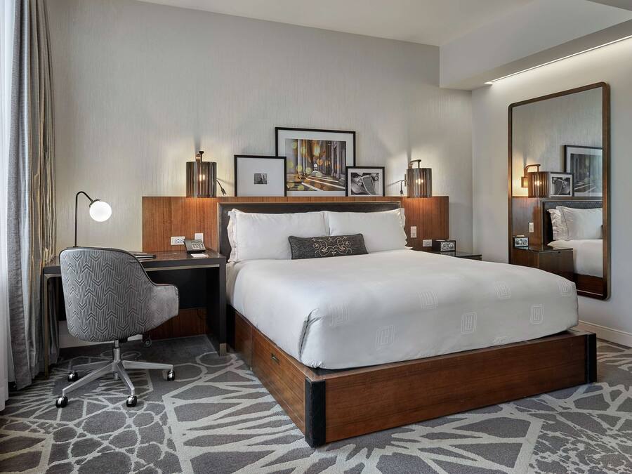A hotel room with a large bed, a desk with a chair, a desk lamp, framed pictures on the wall, and a large mirror.