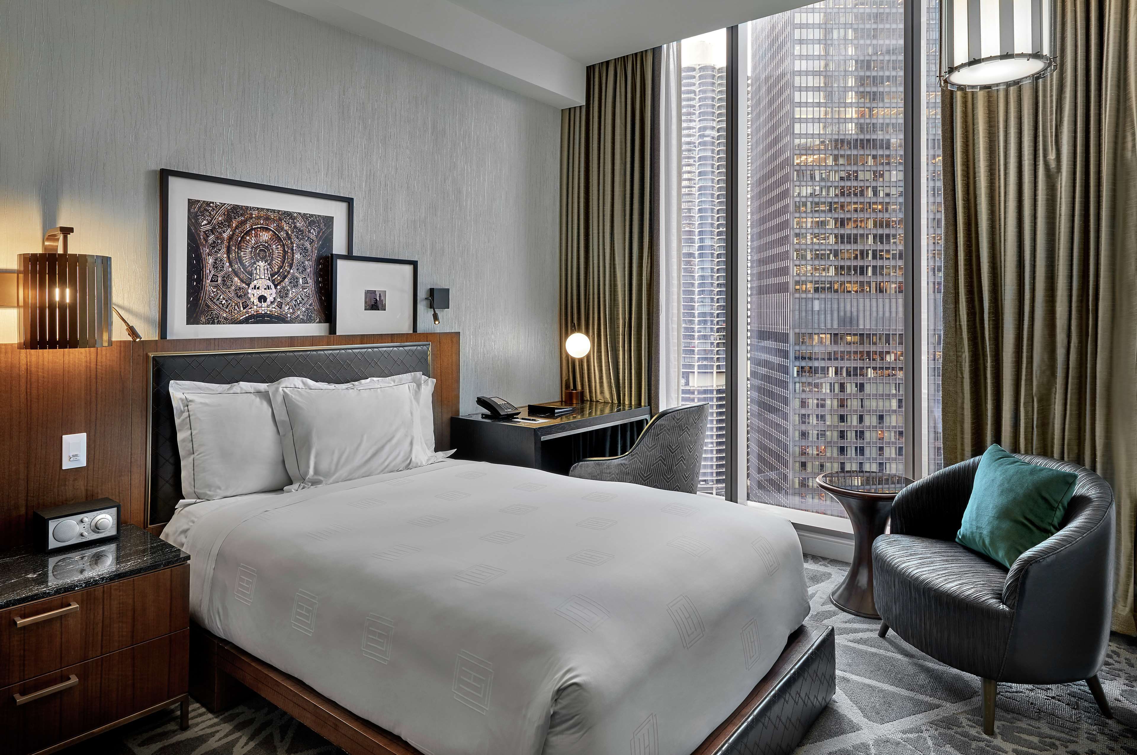 A hotel room with a large bed, a desk, a chair, a lamp, and a view of a cityscape through the window.