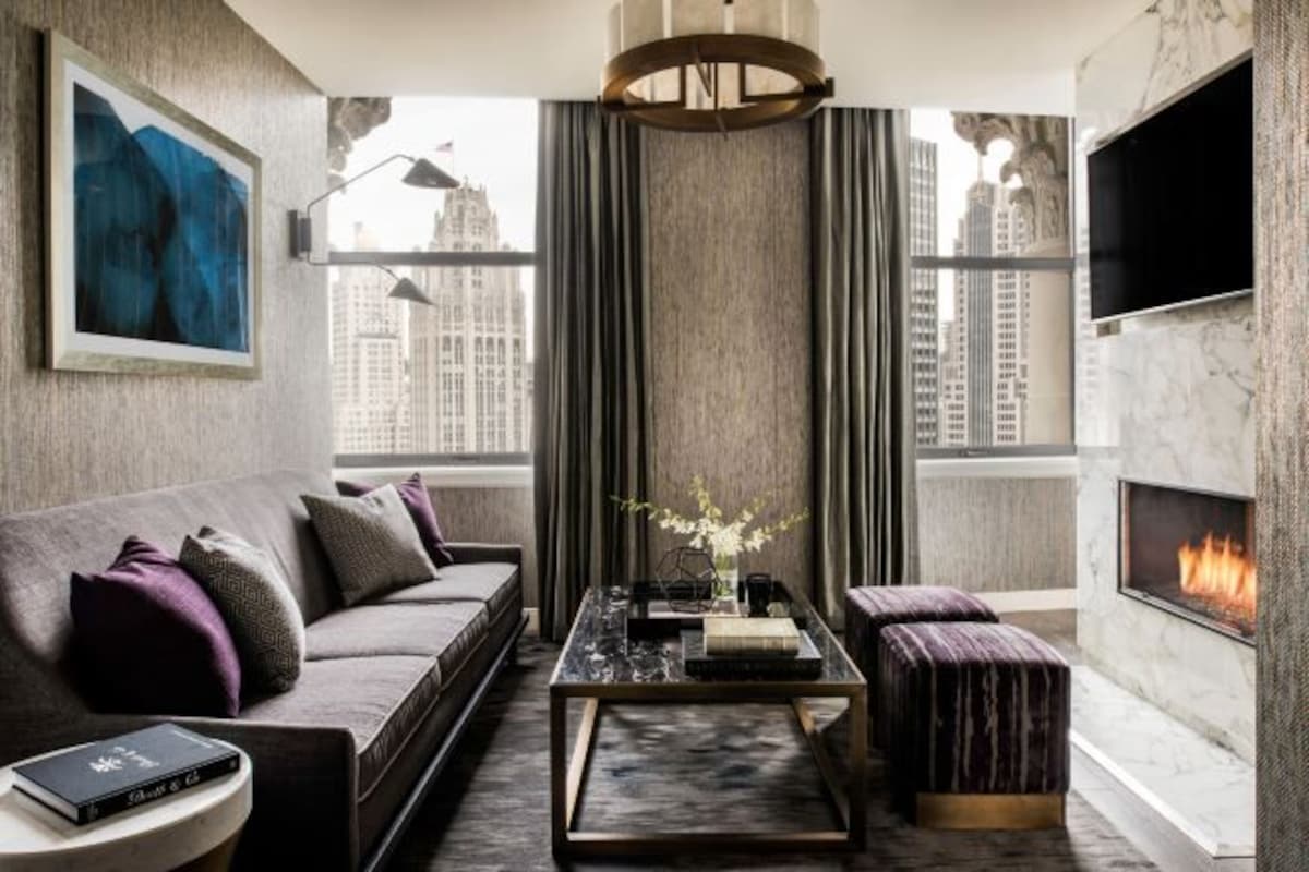 A modern living room with a sofa, coffee table, and ottoman, a fireplace, a TV, and a large window with a city view.