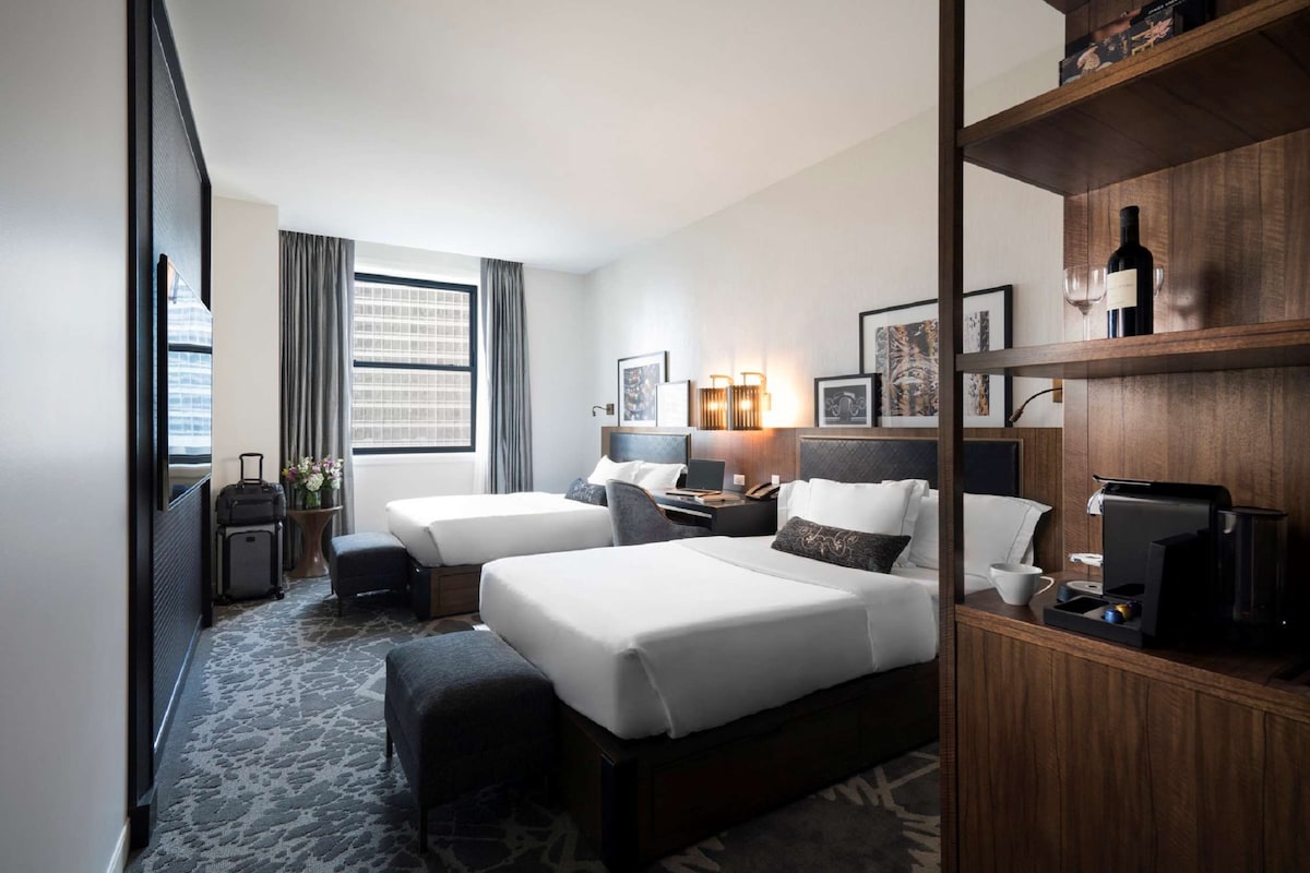 A hotel room with two beds, a desk, a coffee machine, and a bookshelf.