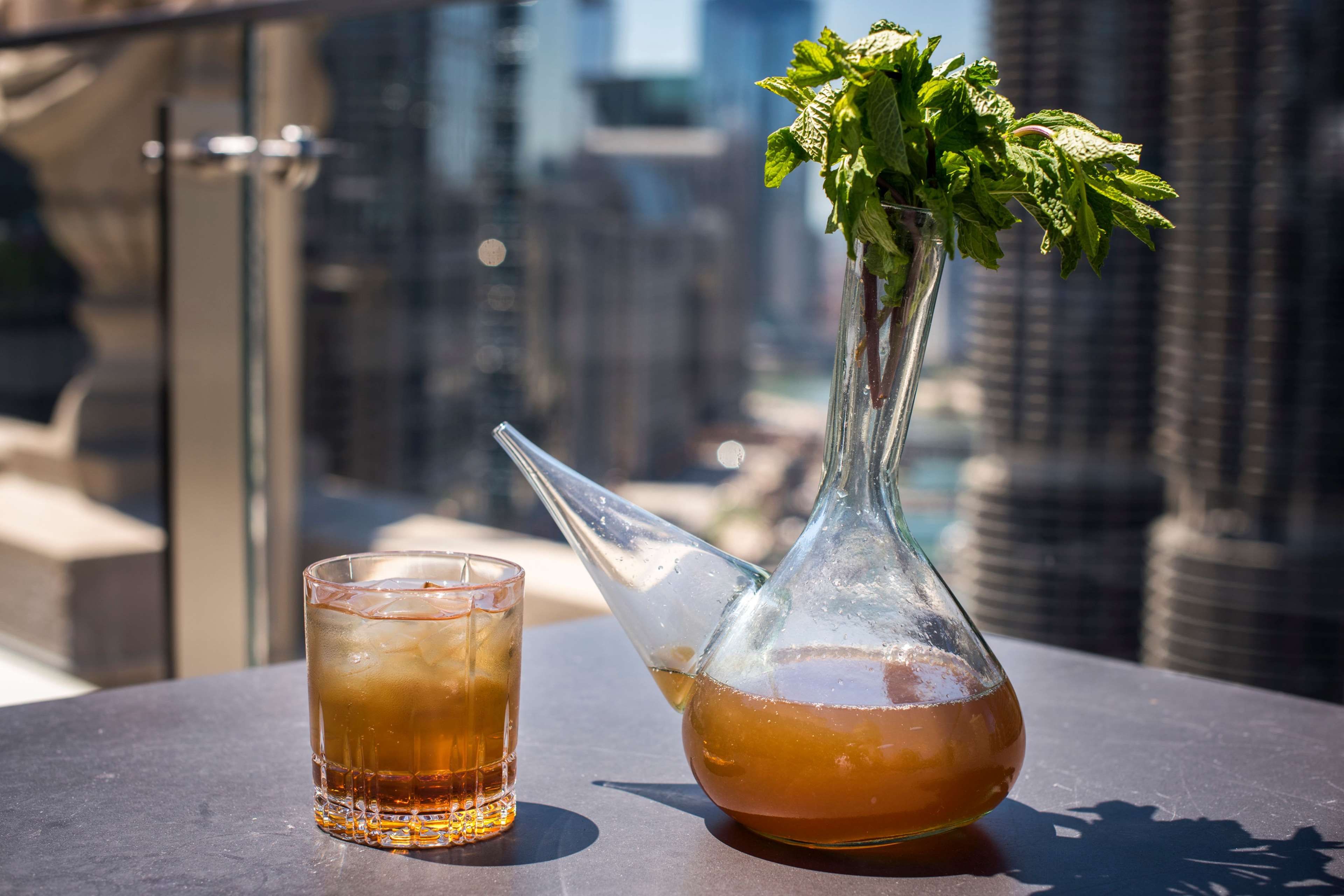 A glass of iced tea and a pitcher with mint leaves on a table with a city view.