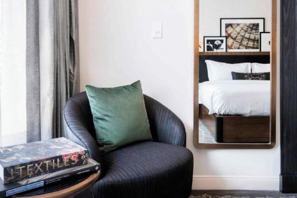 A hotel room with a dark leather armchair, a bedside table with a book, a large mirror reflecting the bed, and framed artwork on the wall.