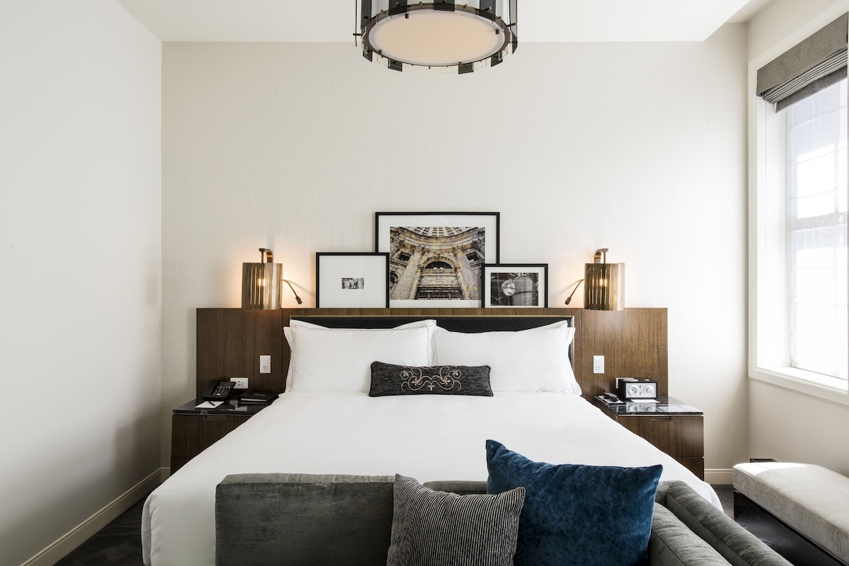 A hotel room with a large bed, two bedside tables with lamps, a window with curtains, and framed pictures on the wall.