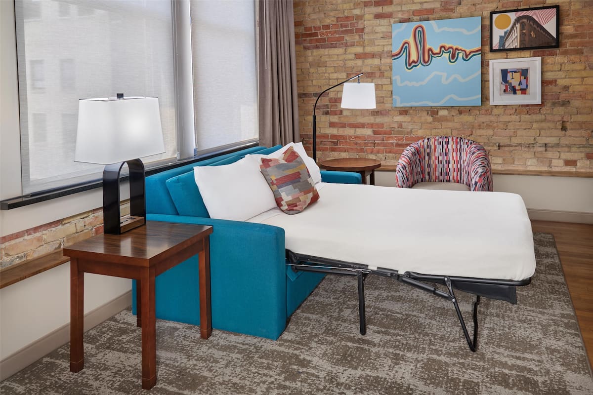 A modern hotel room with a blue sofa, a small wooden table, a foldable bed, a lamp, and artwork on the brick wall.