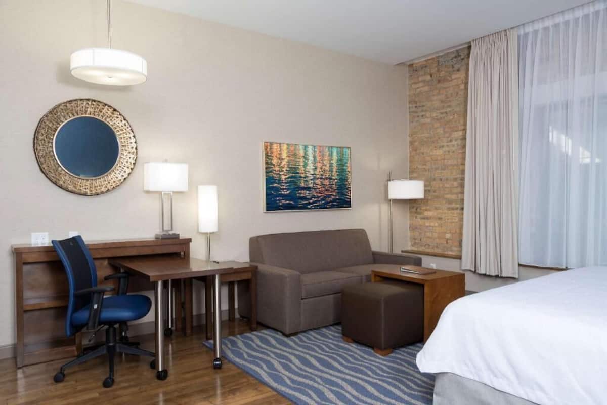 A hotel room with a desk, chair, sofa, ottoman, bed, and artwork on the wall.