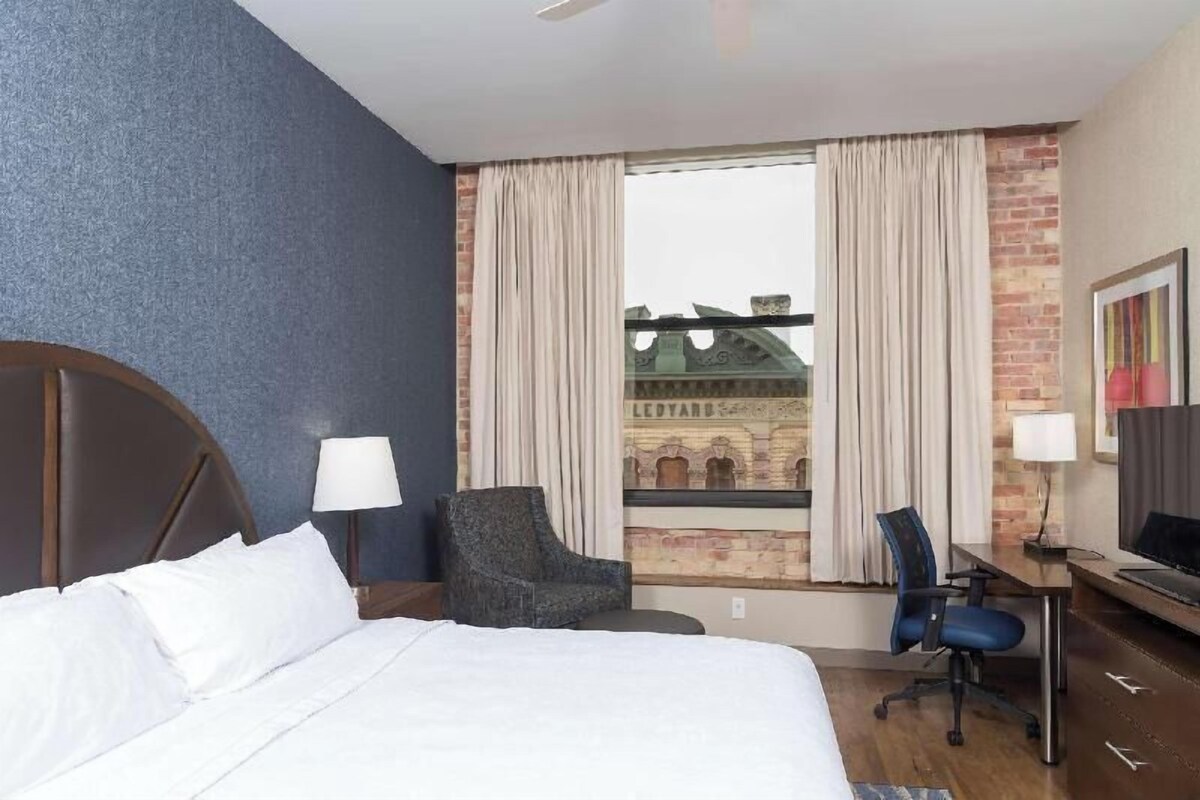 Habitación de hotel con cama, dos sillas, escritorio, televisor y ventana con cortinas.