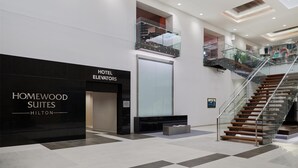 Un moderno vestíbulo de hotel con un letrero negro que dice "Homewood Suites Hilton", un ascensor de vidrio y una escalera con barandillas de cristal.