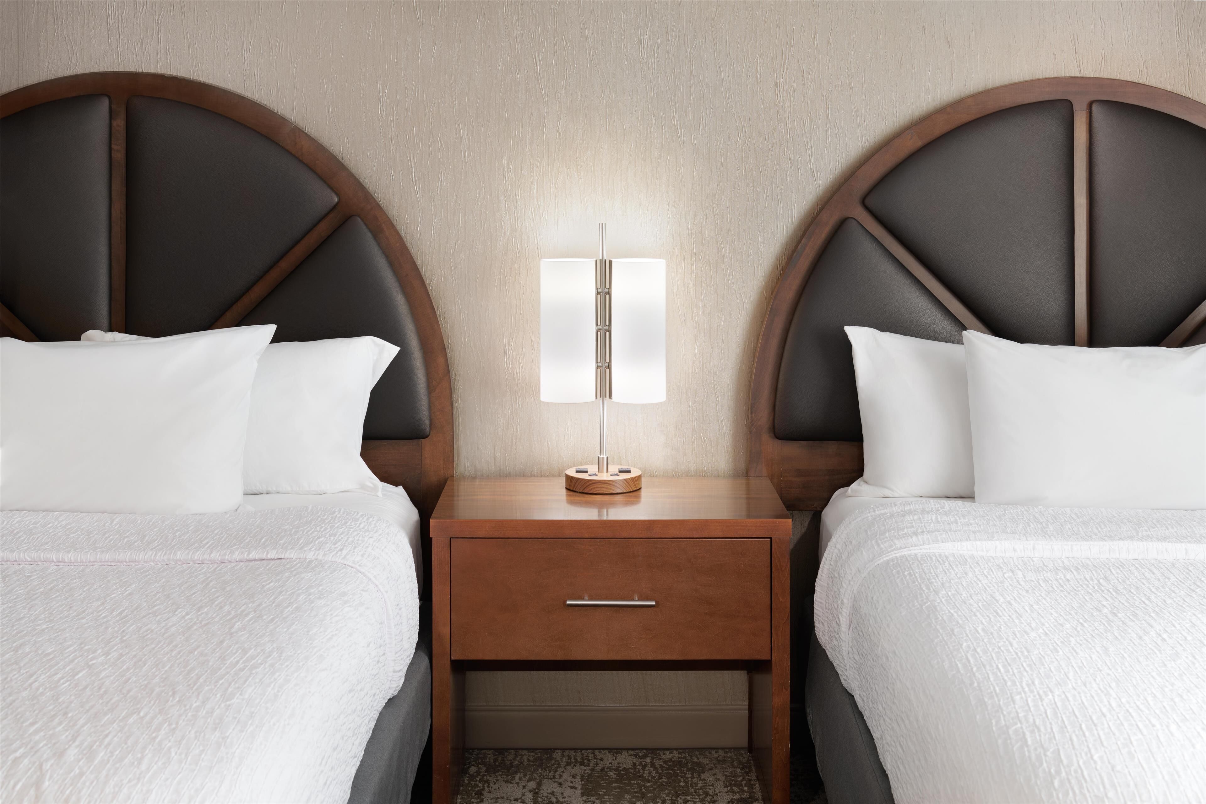 Una habitación de hotel con dos camas, una mesita de noche con lámpara y un cabecero de madera.