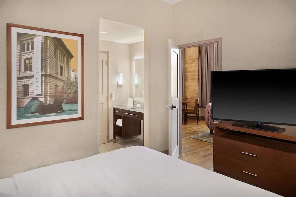 A hotel room with a bed, a television, a desk, and a painting of a building.