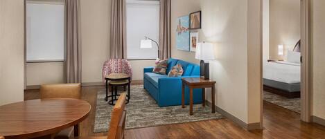 Una habitación de hotel con una mesa de comedor de madera, un sofá azul, un sillón estampado y una cama con colchón blanco.