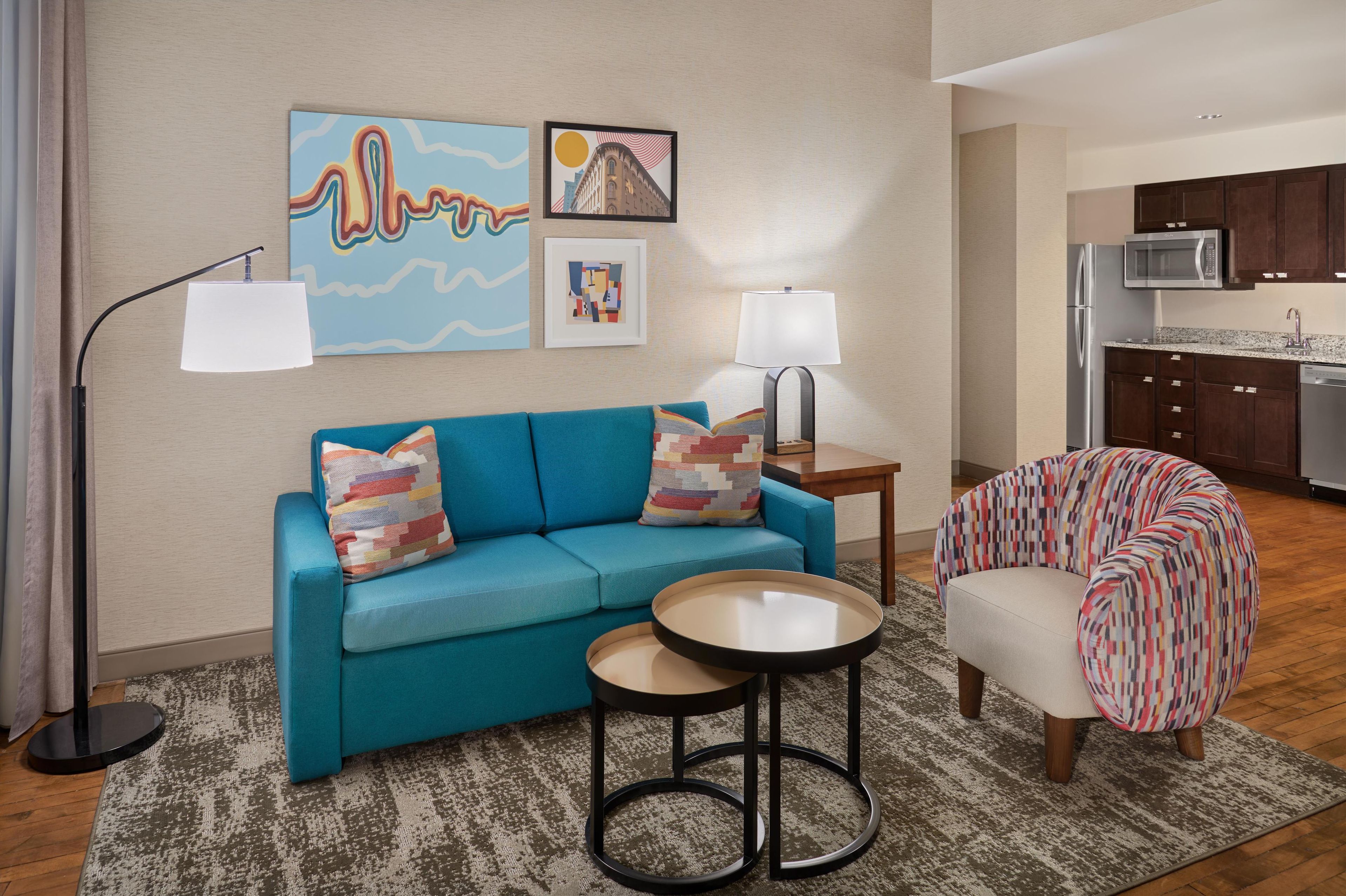 A living room with a blue sofa, two round tables, a patterned armchair, and a kitchen in the background.