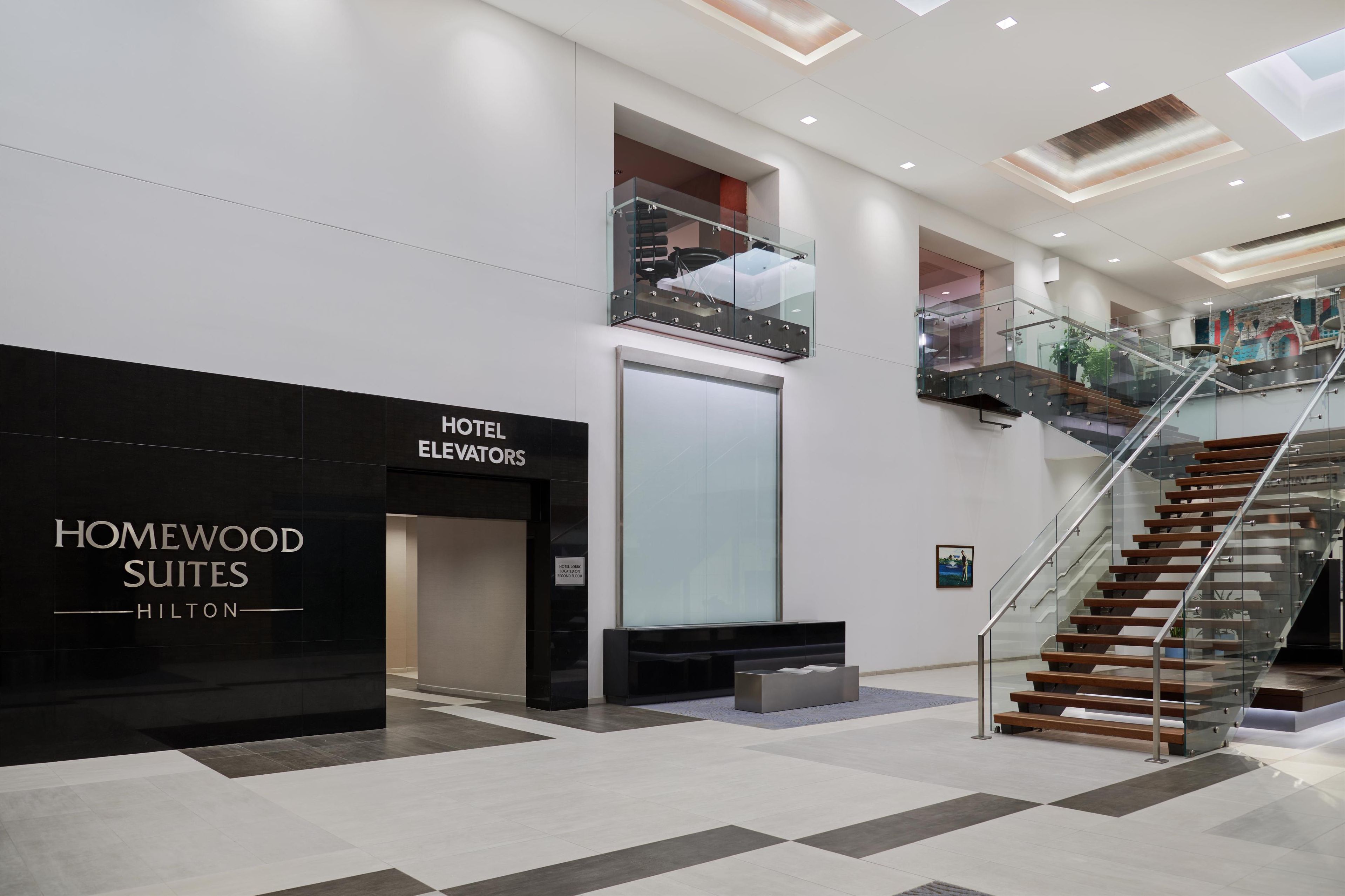 A modern hotel lobby with a black "Homewood Suites Hilton" sign, a glass elevator, and a staircase with glass railings.