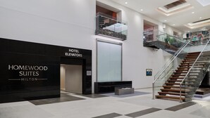 Un moderno vestíbulo de hotel con un letrero negro que dice "Homewood Suites Hilton", un ascensor de vidrio y una escalera con barandillas de cristal.