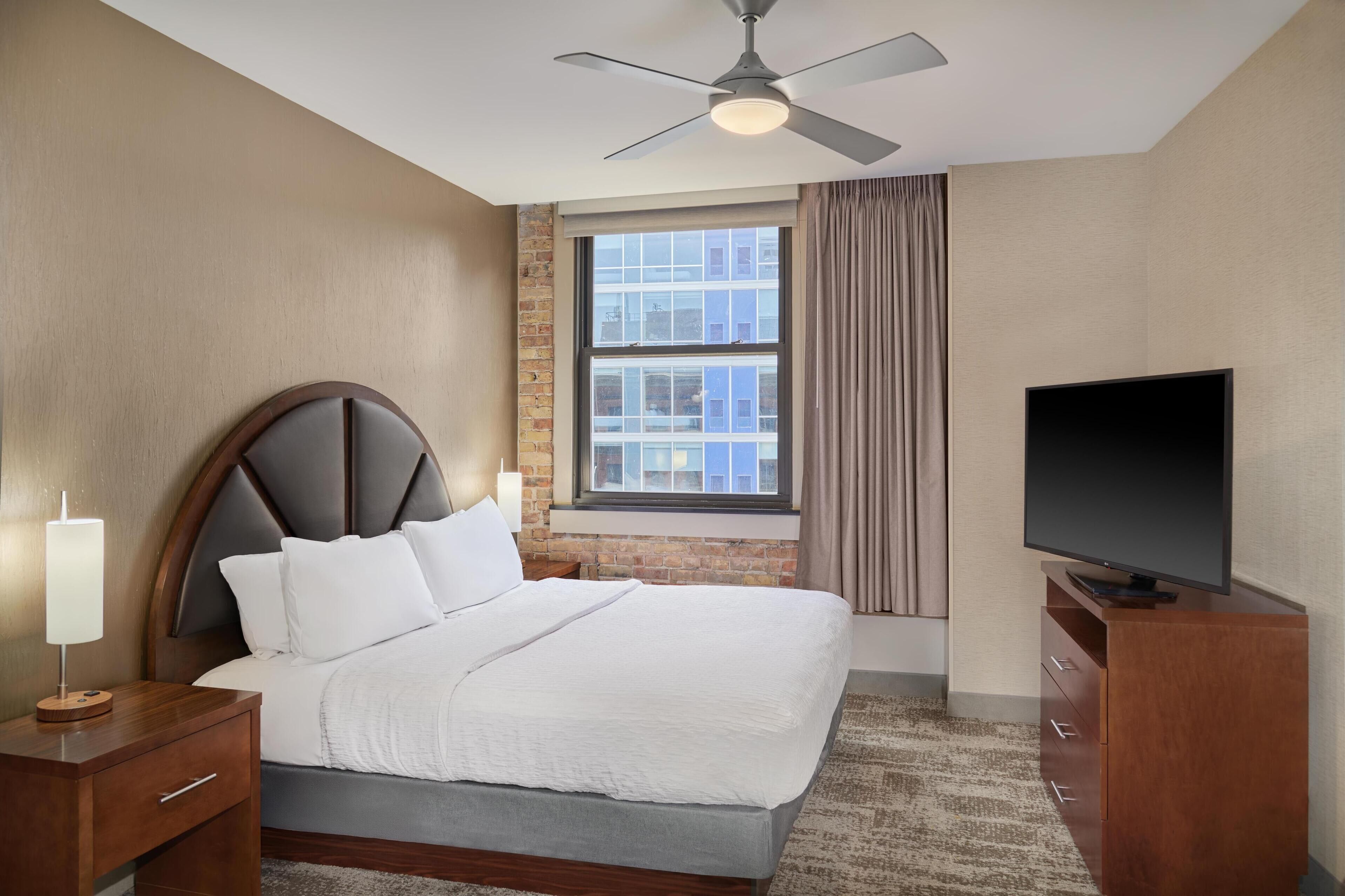 Habitación de hotel con cama, televisión, mesita de noche, lámpara, ventana con cortinas y ventilador de techo.