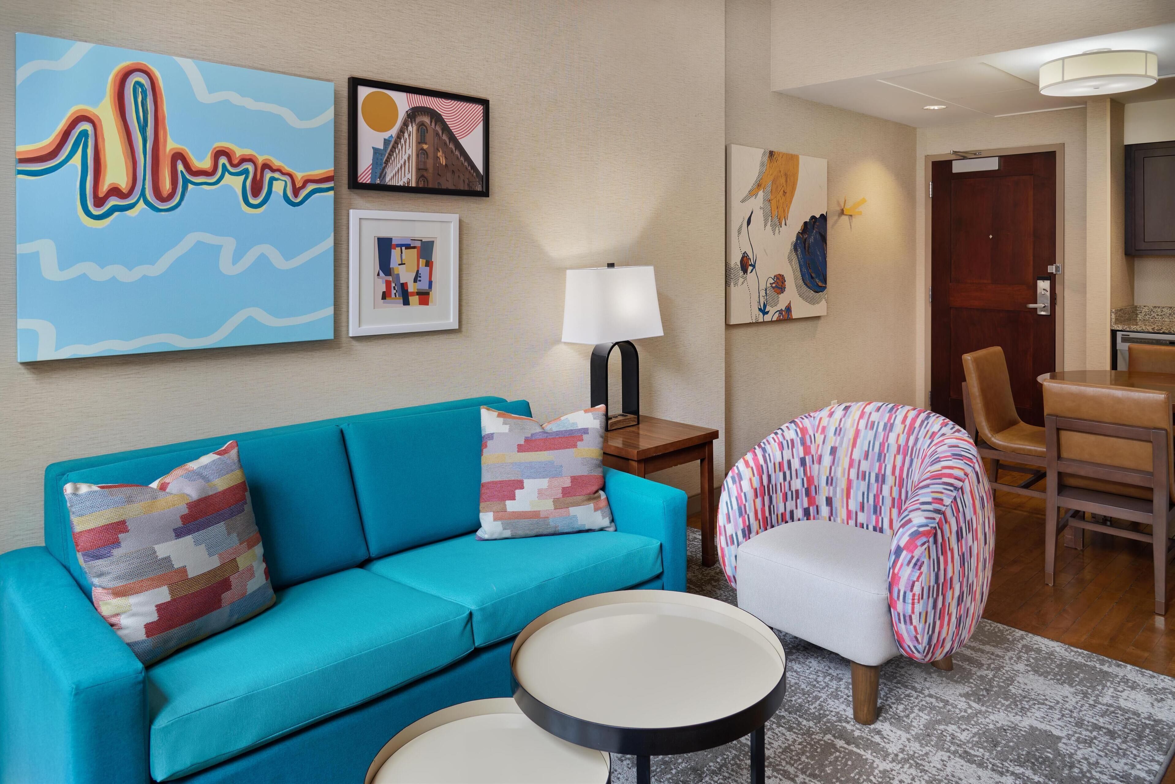Una sala de estar moderna con un sofá azul, una mesa de centro redonda y un sillón de colores. Se observan obras de arte enmarcadas en la pared y una lámpara sobre una mesita auxiliar.