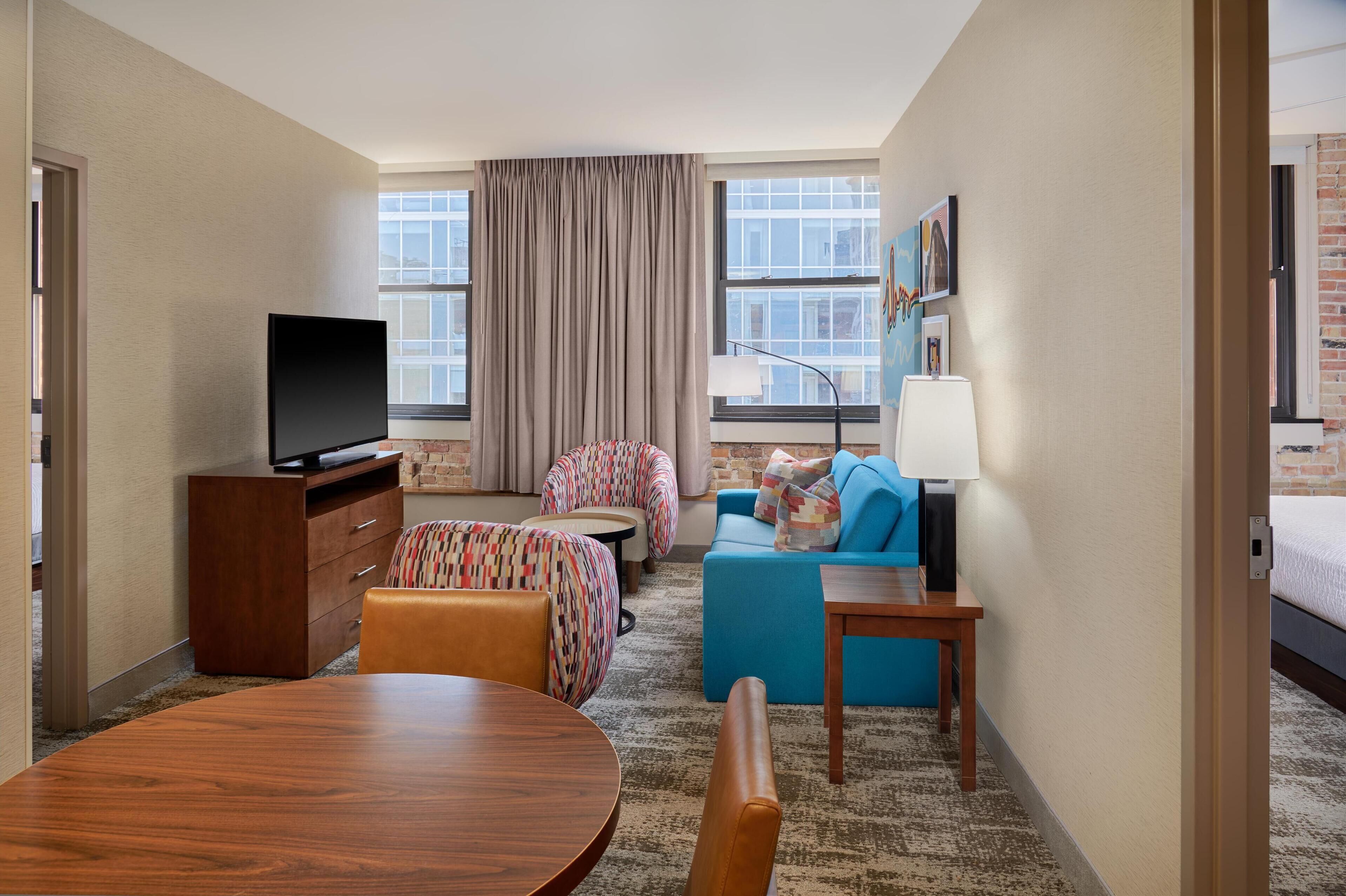 Ein Hotelzimmer mit Fernseher, Esstisch, Sofa, einem kleinen runden Tisch und einer Lampe.