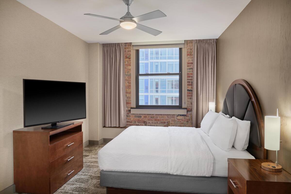 A hotel room with a flat-screen TV, a bed with white linens, a wooden nightstand, and a ceiling fan.
