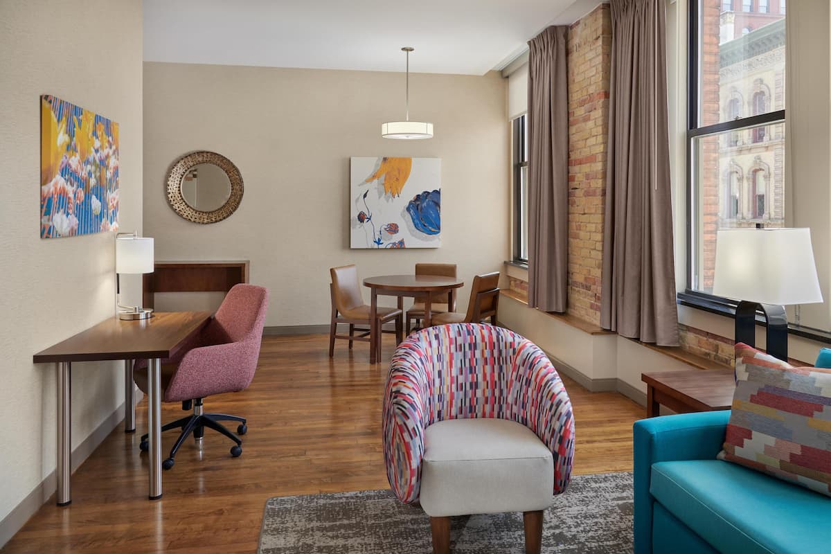 A modern hotel room with a wooden floor, a round table, a sofa, a chair, and a bed.