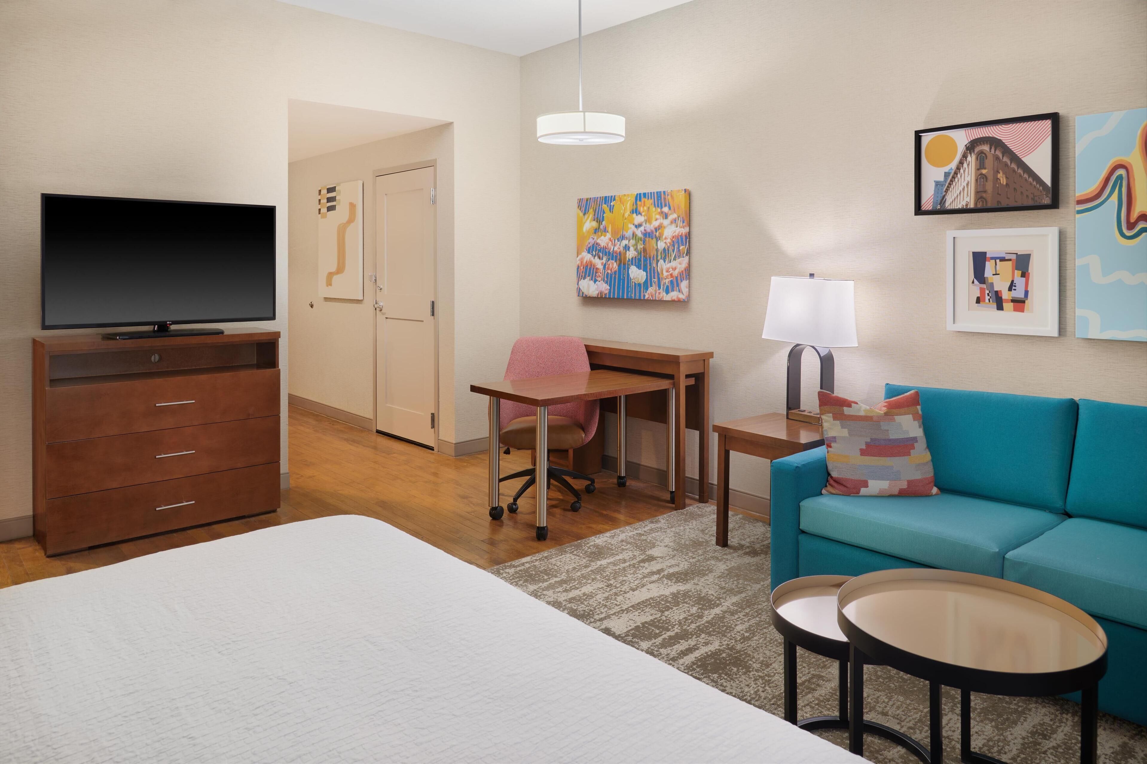 A hotel room with a bed, a desk, a chair, a TV, a sofa, two side tables, a lamp, and artwork on the walls.