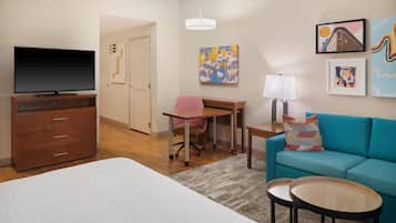 A hotel room with a bed, a desk, a chair, a TV, a sofa, two side tables, a lamp, and artwork on the walls.