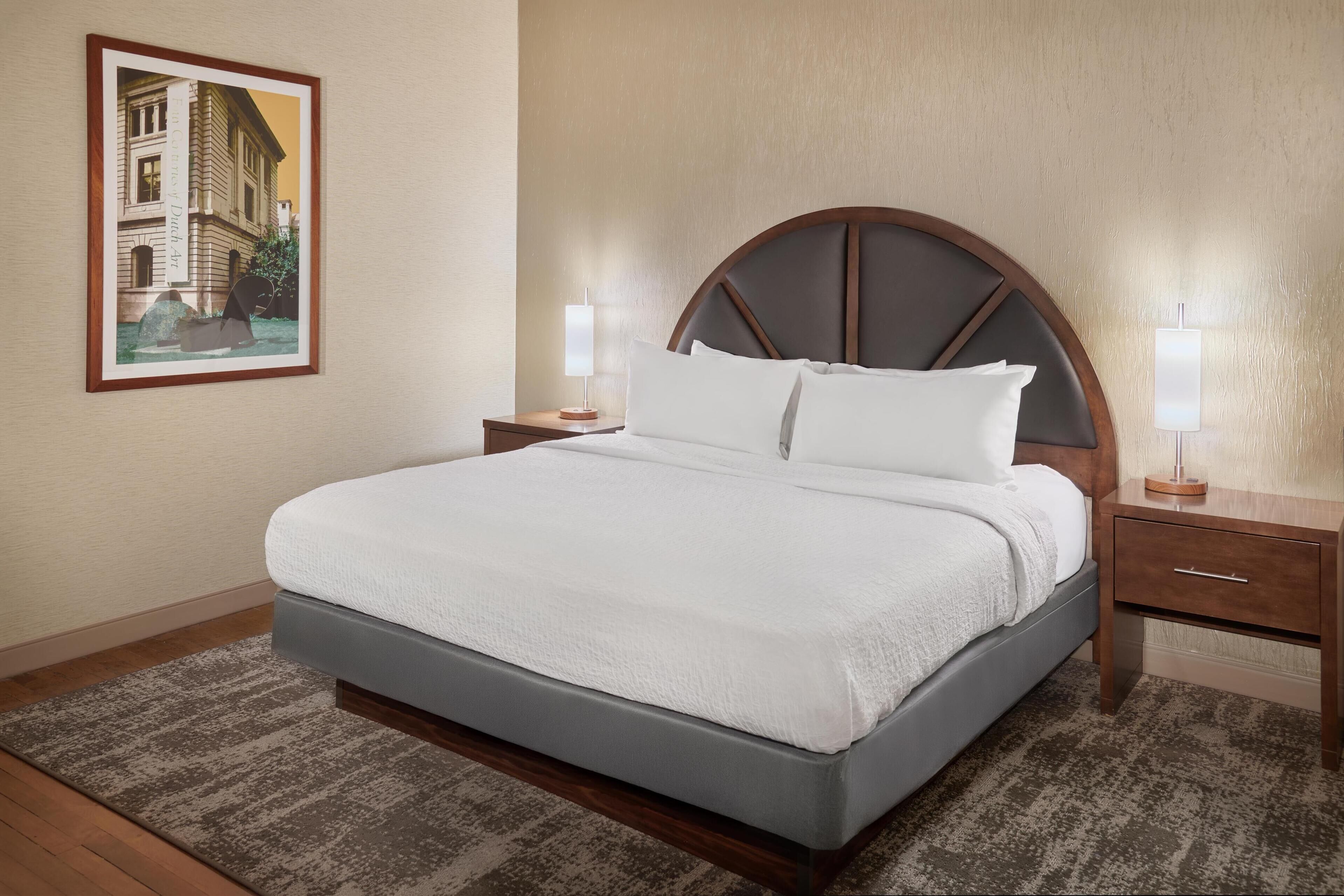 Una cama bien tendida con cabecera de madera, dos mesitas de noche con lámparas y un cuadro en la pared.