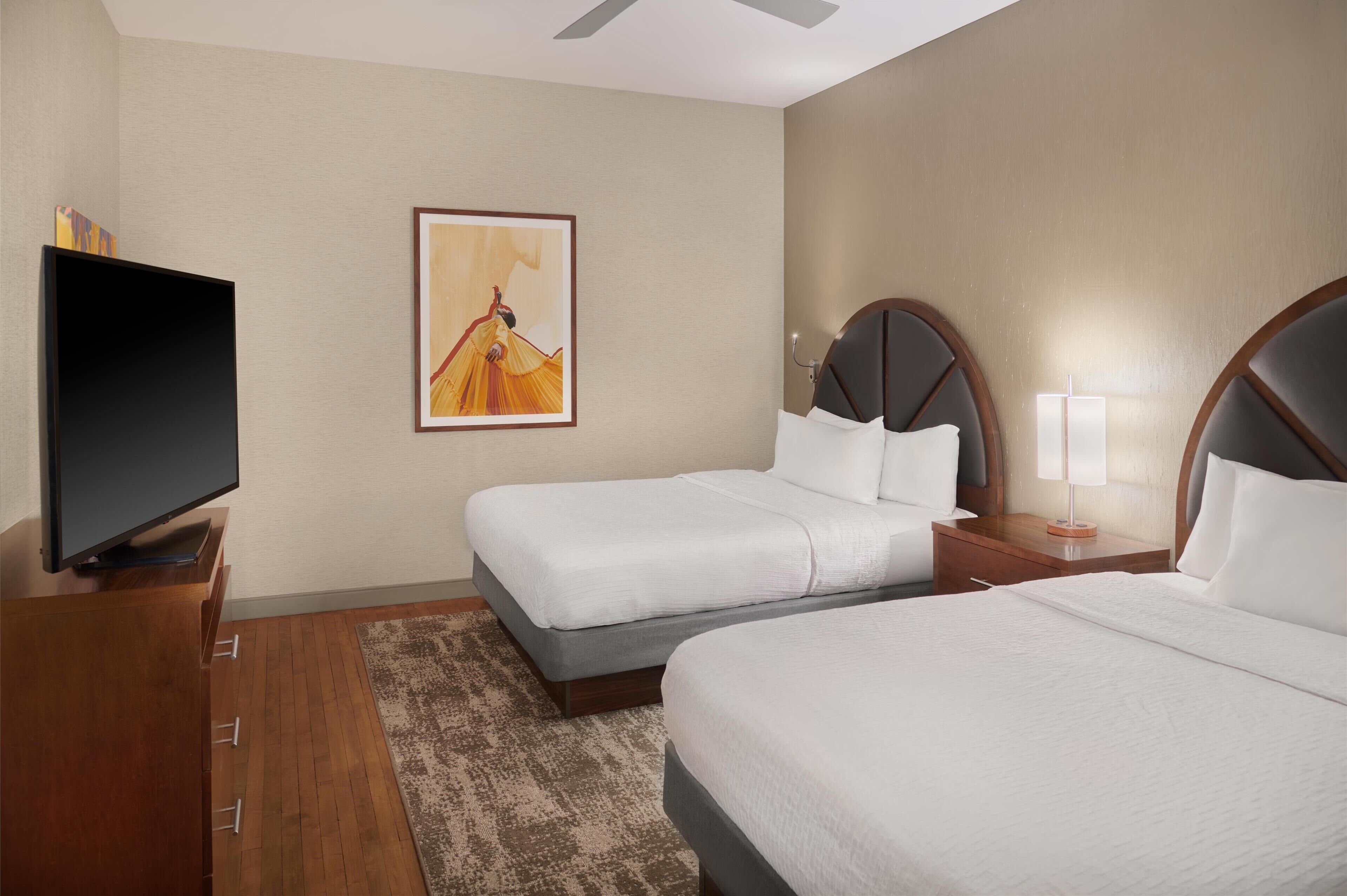 A hotel room with two beds, a TV, a painting, and a lamp.