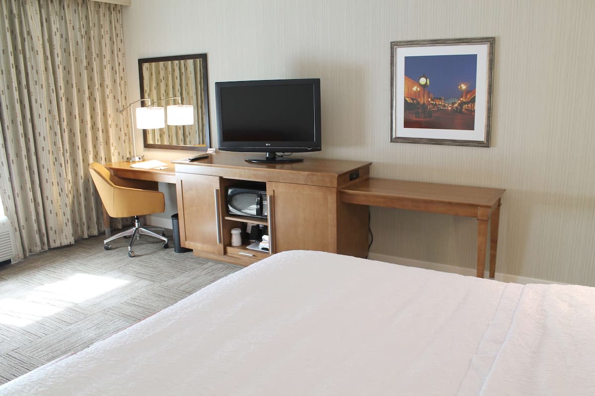 A hotel room with a bed, a desk with a computer, a chair, a lamp, and a framed picture on the wall.