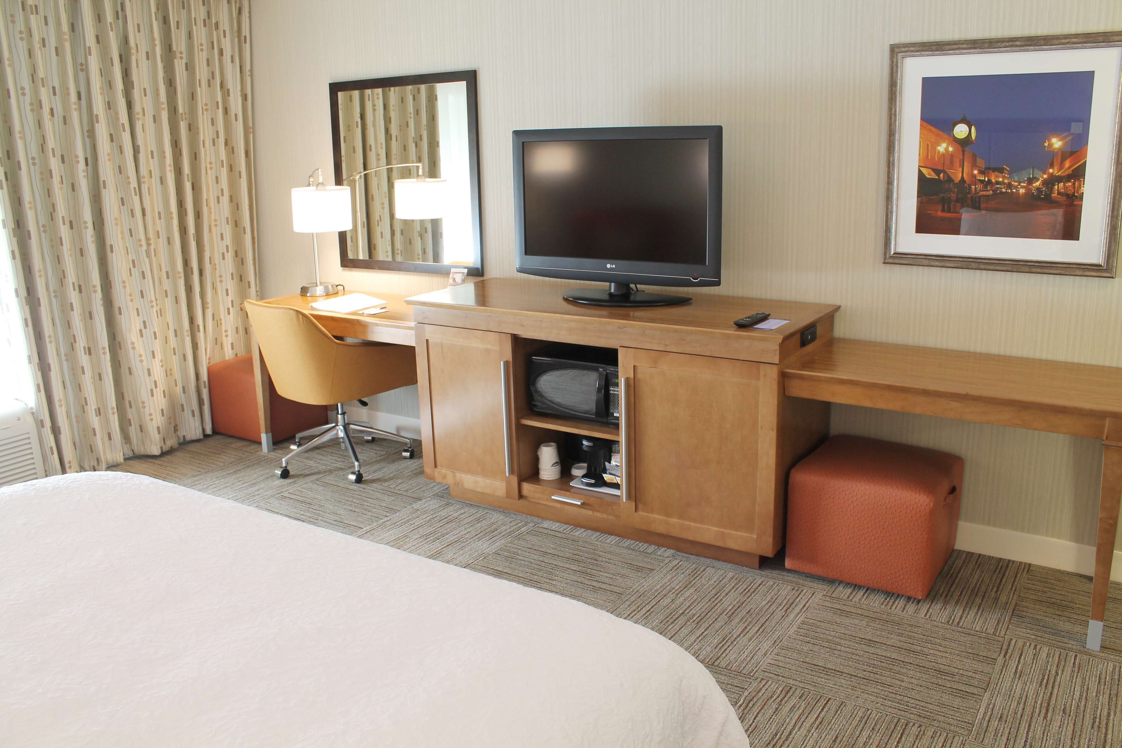 A hotel room with a bed, a desk with a computer, a chair, a lamp, a television, a framed picture on the wall, and a carpeted floor.