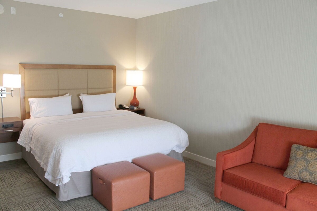 A hotel room with a bed, a nightstand with a lamp, a red sofa, and a small ottoman.