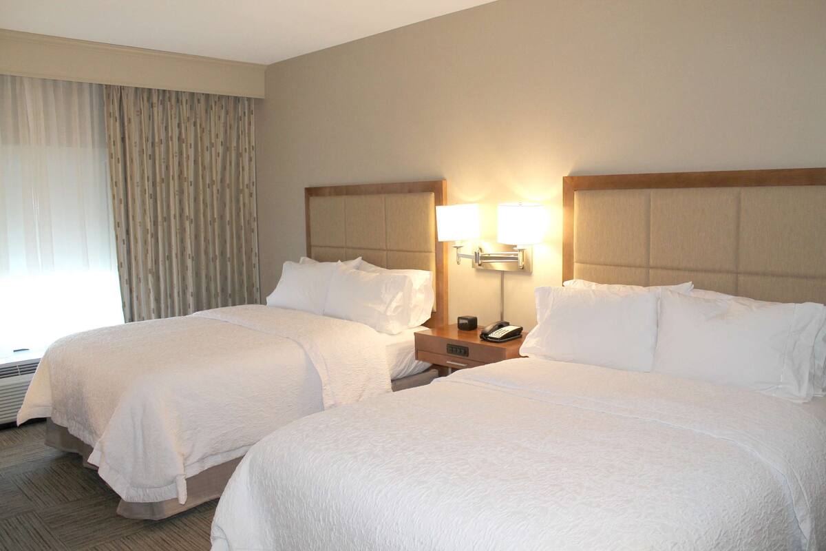 A hotel room with two beds, a nightstand with a lamp, and a window with curtains.