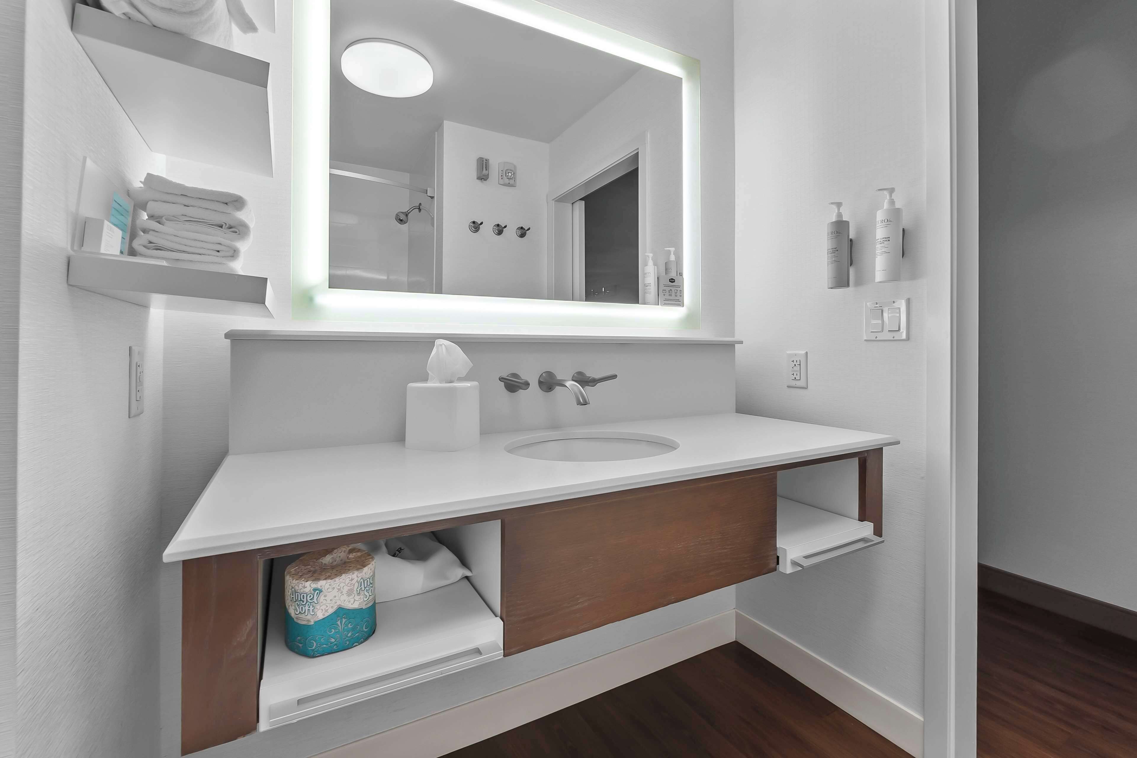 A modern bathroom with a white sink, wooden vanity, and a large mirror with integrated lighting.