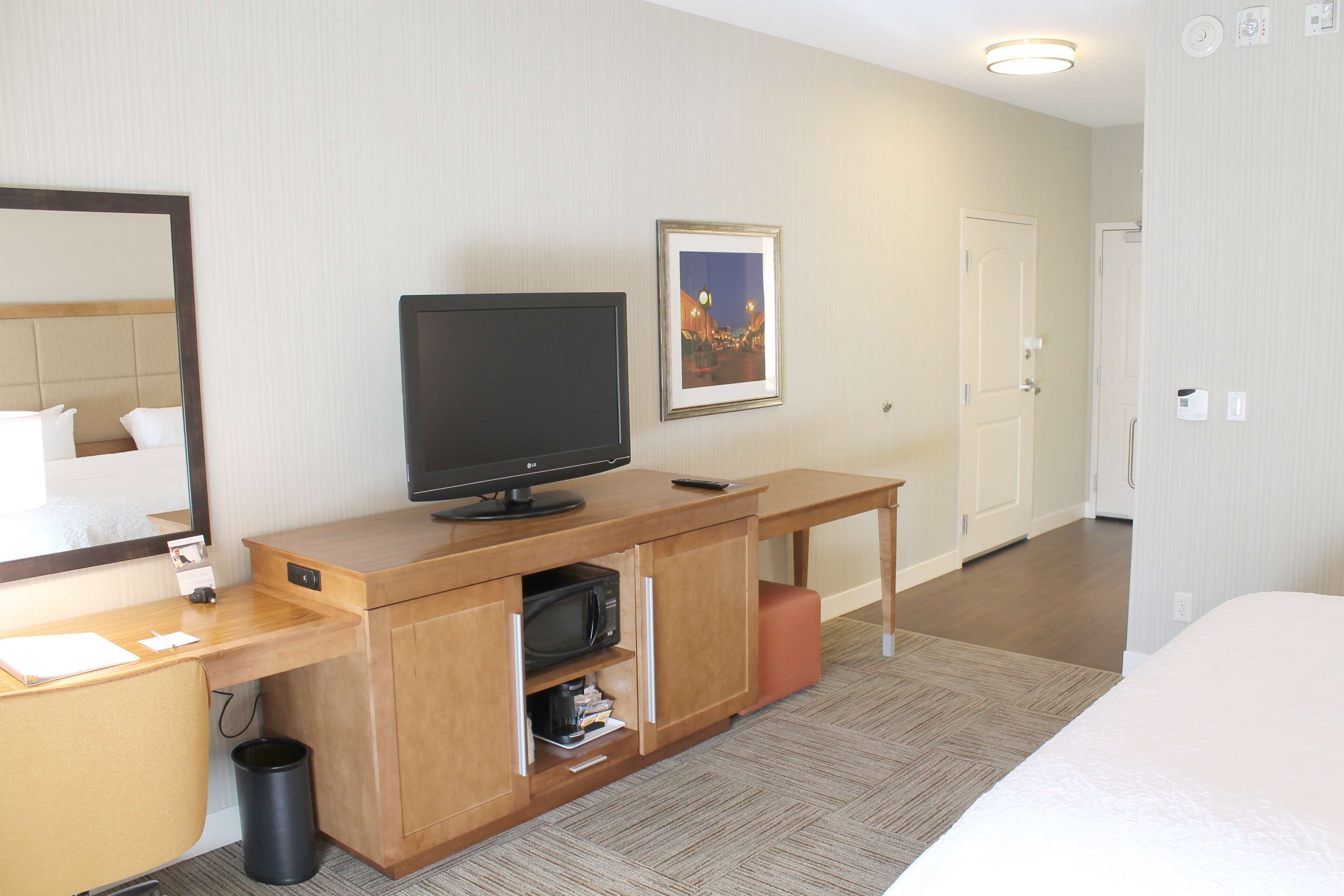 A hotel room with a wooden desk, a flat-screen TV, a framed picture on the wall, and a bed.