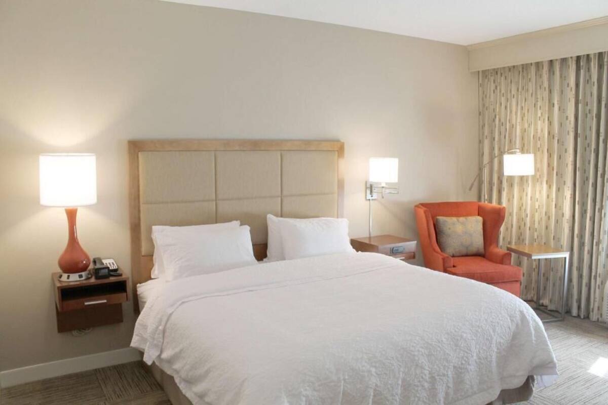 A hotel room with a large bed, two bedside lamps, a nightstand with a phone, a chair, and a curtain.